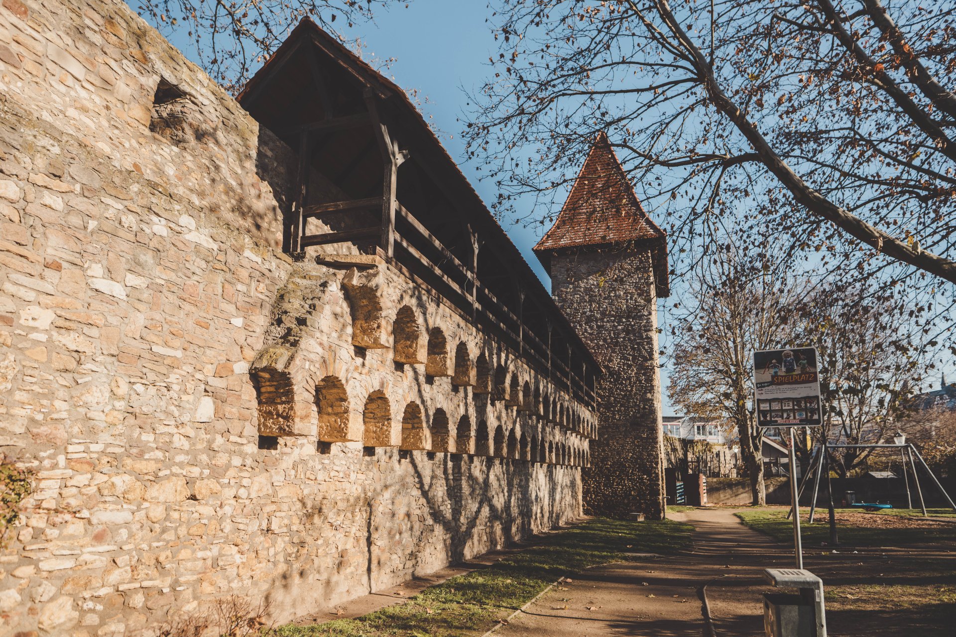 Historische Stadtmauer mit kleinen Bögen und einem steinernen Turm, umgeben von hohen, kahlen Bäumen.