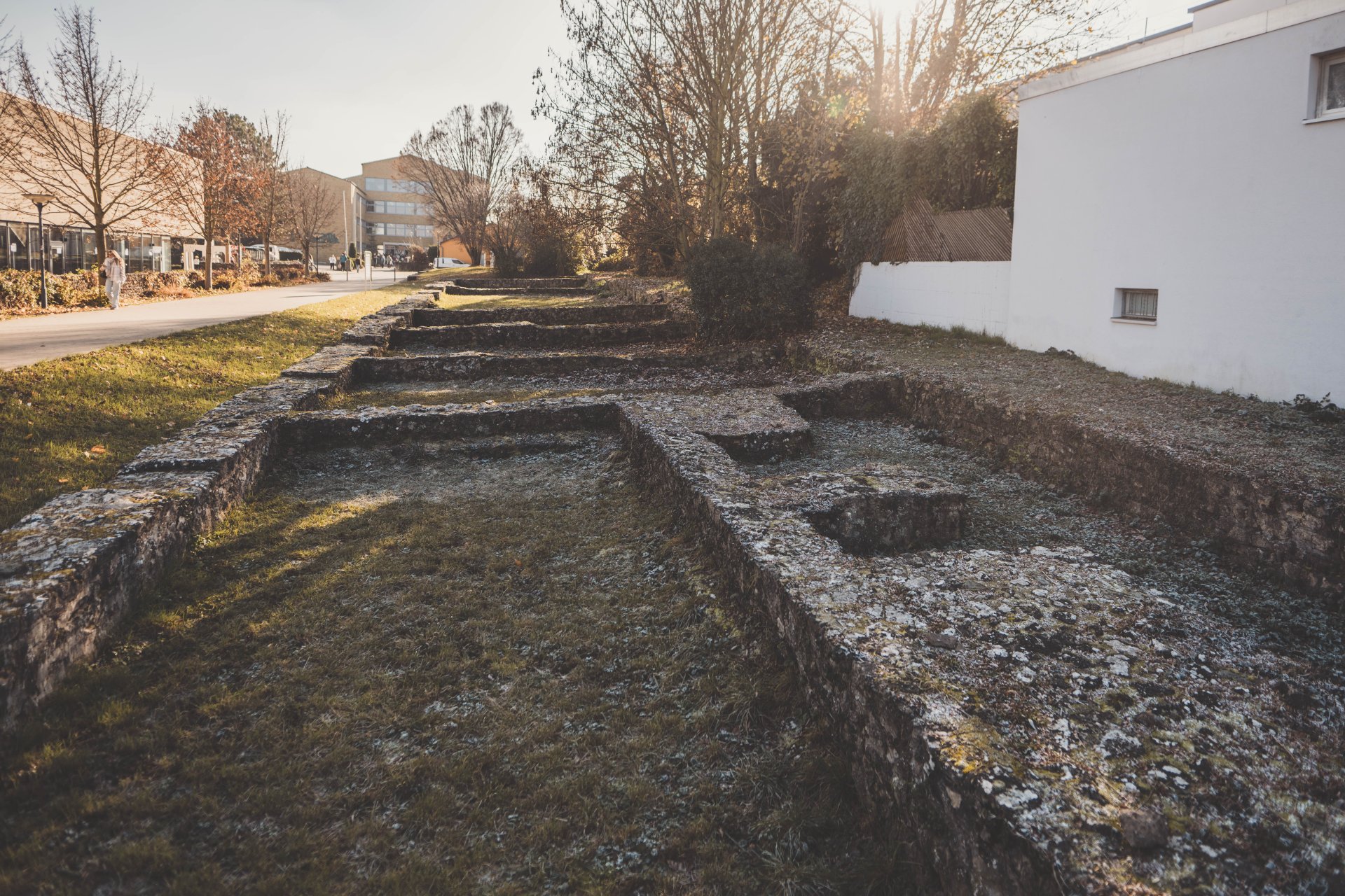 Freigelegte Mauern des ehemaligen Römerkastells im Vordergrund, dahinter moderne Gebäude und herbstliche Bäume im warmen Sonnenlicht.