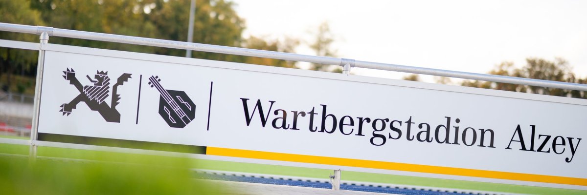 Zu sehen ist ein Schild mit der Aufschrift „Wartbergstadion Alzey“. Das Schild steht am Rand eines Sportplatzes, im Hintergrund sind Bäume und ein Stadionbereich zu erkennen.