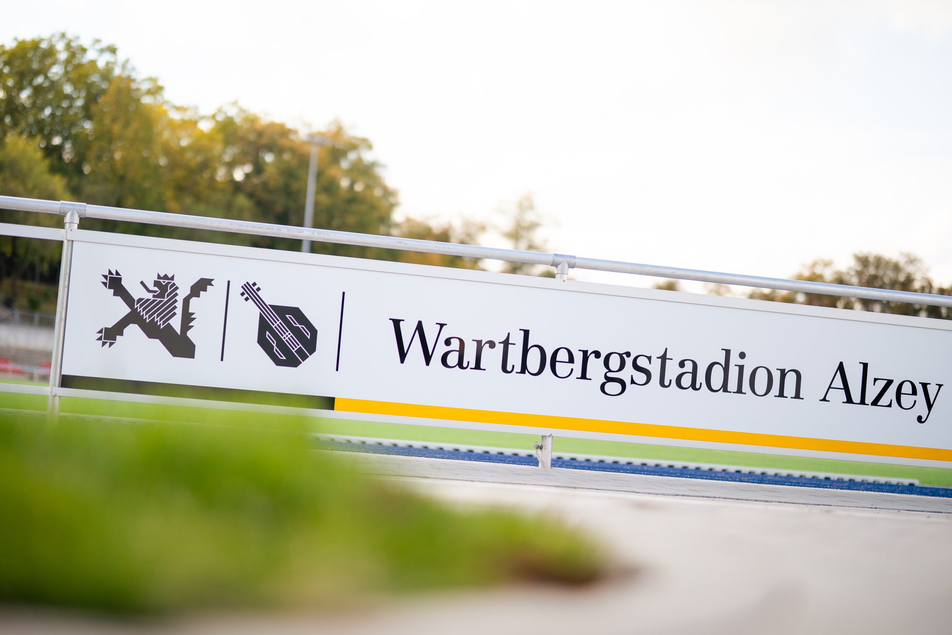 Zu sehen ist ein Schild mit der Aufschrift „Wartbergstadion Alzey“. Das Schild steht am Rand eines Sportplatzes, im Hintergrund sind Bäume und ein Stadionbereich zu erkennen.