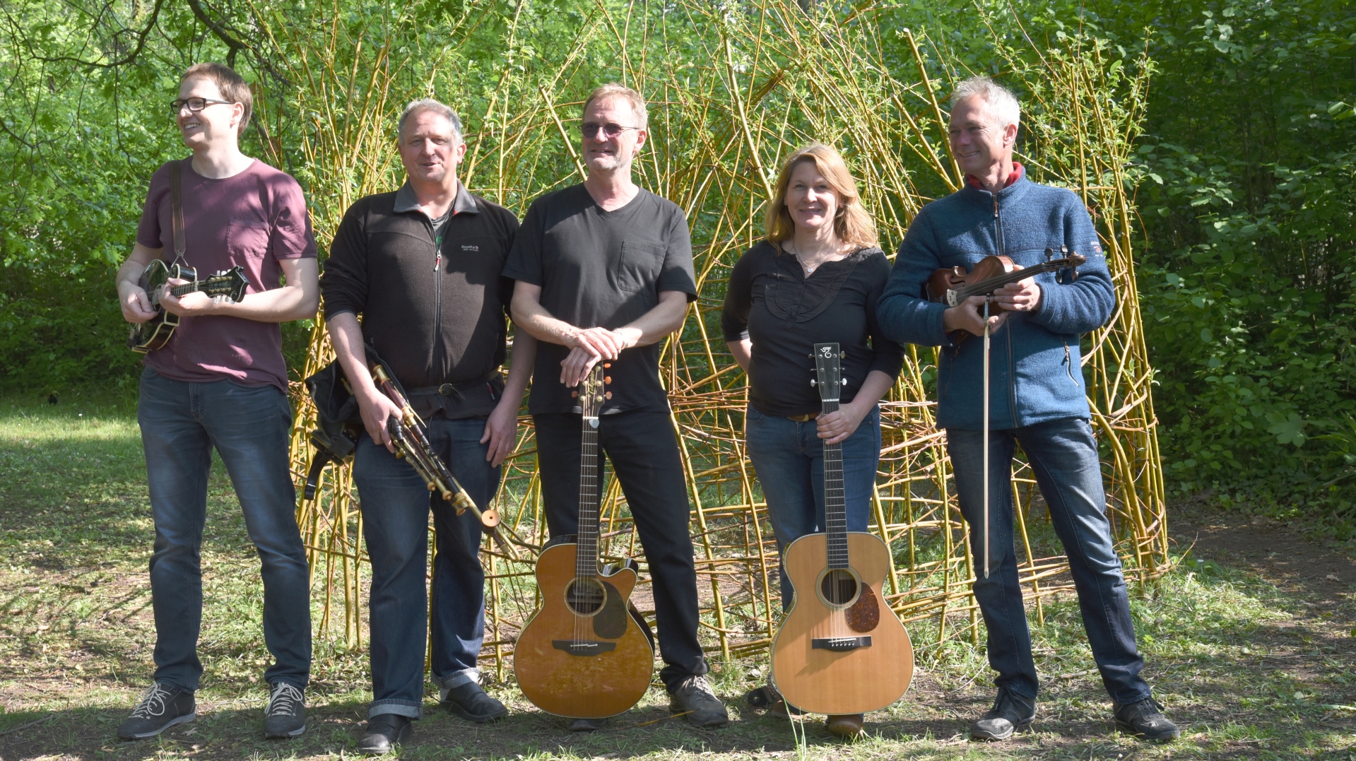 Man sieht die Gruppe Fleadh im Wald auf einer Lichtung stehend mit ihren Musikinstrumenten in die Kamera schauend.