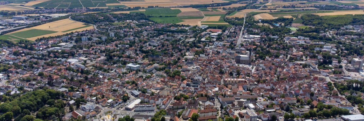 Die Stadt Alzey aus einer schrägen Luftansicht. Hinter der Stadt liegen Felder und Weinberge mit Windrädern. Im Vordergrund zu sehen, ist der Bahnhof und das angrenzende Neubaugebiet.