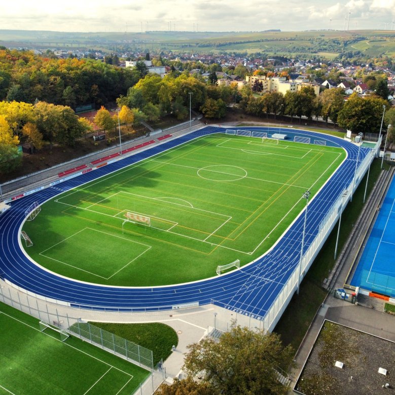 Die neuen Sportplätze des Wartbergstadions. Um den grünen Rasenplatz, zieht sich eine dunkelblaue Tartanbahn. Auf der langen Seite der Bahn befinden sich mehrere Reihen roter Sitzschalen. Rechts daneben, direkt unterhalb des Platzes befindet sich in einem helleren Blau der erneuerte Hockeyplatz. Im Hintergrund sieht man einen Teil der Stadt Alzey.