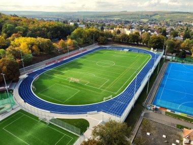 Die neuen Sportplätze des Wartbergstadions. Um den grünen Rasenplatz, zieht sich eine dunkelblaue Tartanbahn. Auf der langen Seite der Bahn befinden sich mehrere Reihen roter Sitzschalen. Rechts daneben, direkt unterhalb des Platzes befindet sich in einem helleren Blau der erneuerte Hockeyplatz. Im Hintergrund sieht man einen Teil der Stadt Alzey.