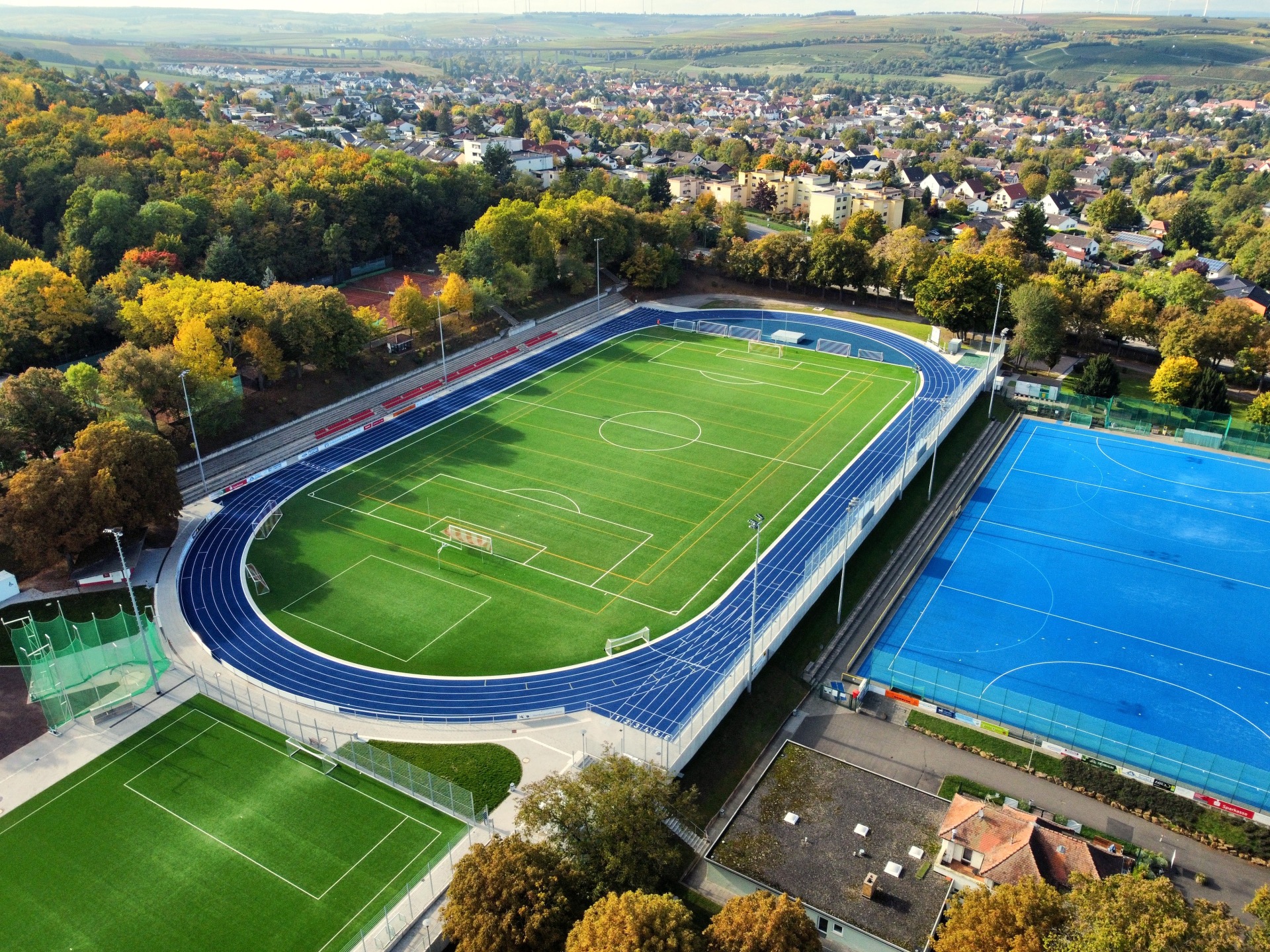 Die neuen Sportplätze des Wartbergstadions. Um den grünen Rasenplatz, zieht sich eine dunkelblaue Tartanbahn. Auf der langen Seite der Bahn befinden sich mehrere Reihen roter Sitzschalen. Rechts daneben, direkt unterhalb des Platzes befindet sich in einem helleren Blau der erneuerte Hockeyplatz. Im Hintergrund sieht man einen Teil der Stadt Alzey.