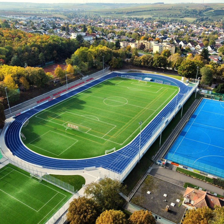 Die neuen Sportplätze des Wartbergstadions. Um den grünen Rasenplatz, zieht sich eine dunkelblaue Tartanbahn. Auf der langen Seite der Bahn befinden sich mehrere Reihen roter Sitzschalen. Rechts daneben, direkt unterhalb des Platzes befindet sich in einem helleren Blau der erneuerte Hockeyplatz. Im Hintergrund sieht man einen Teil der Stadt Alzey.