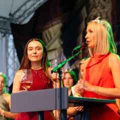 Zwei Frauen stehen auf einer Bühne, die eine spricht ins Mikrofon, die andere hört ihr daneben zu.