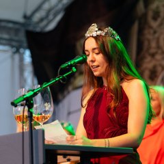 Eine Frau in einem dunkel-rote Kleid trägt eine Krone und spricht auf einer Bühne in ein Mikrofon.