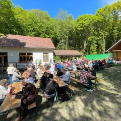 mehrere Personen sitzen auf Bierzeltgarnituren auf einer Wiese zwischen Hütten und essen, einige trinken ein Glas Wein