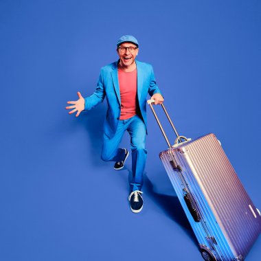 ein Mann in blauem Anzug und einem Koffer in der Hand schaut aufgeregt und lachend in die Kamera