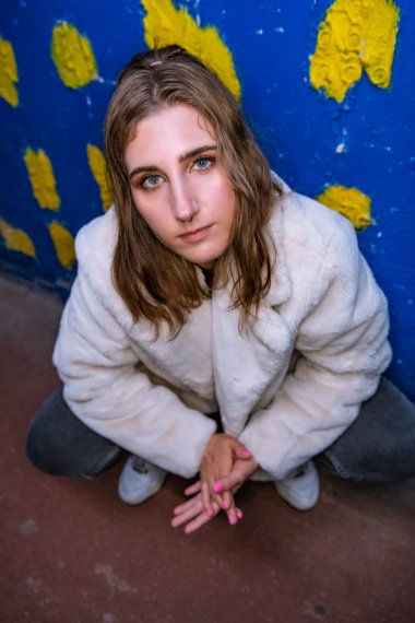 Eine junge Frau sitzt in der Hocke und wird von oben herab fotografiert. Hinter ihr ist eine blaue Wand mit gelben Punkten.