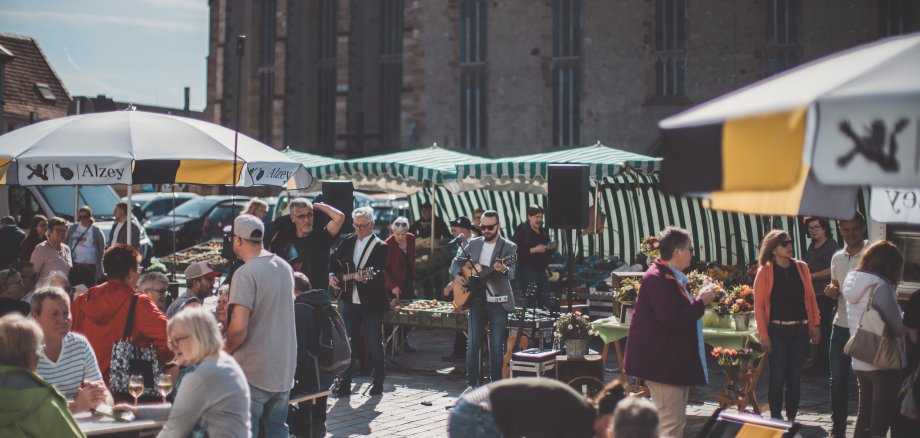 Zwischen den Marktständen sitzen die Leute unter Schirmen auf Bierzeltgarnituren. Zwei Musiker mit Gitarren und Sonnenbrillen stehen in der Mitte.
