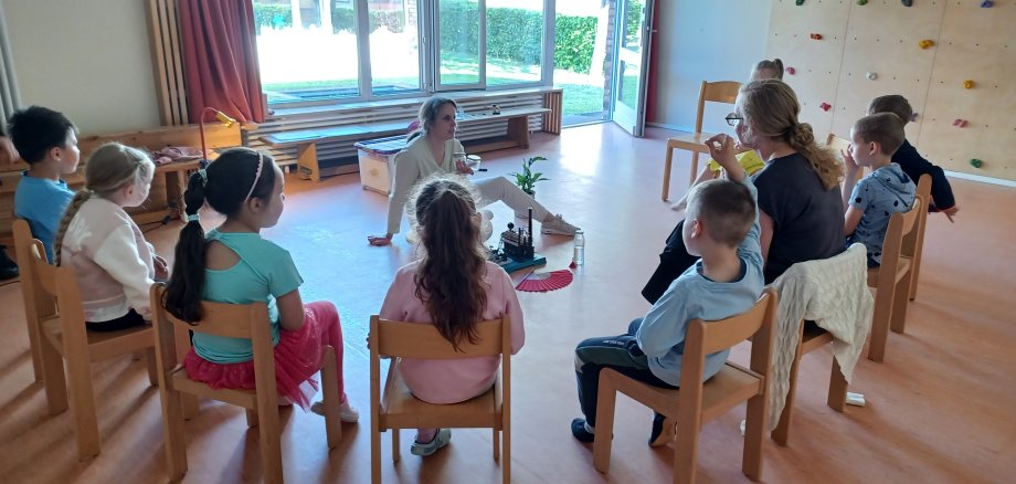 In einem Stuhlkreis sitzen die Kinder und beobachten die Frau in der Mitte dabei, wie sie eine kleine Dampfmaschine anwirft.