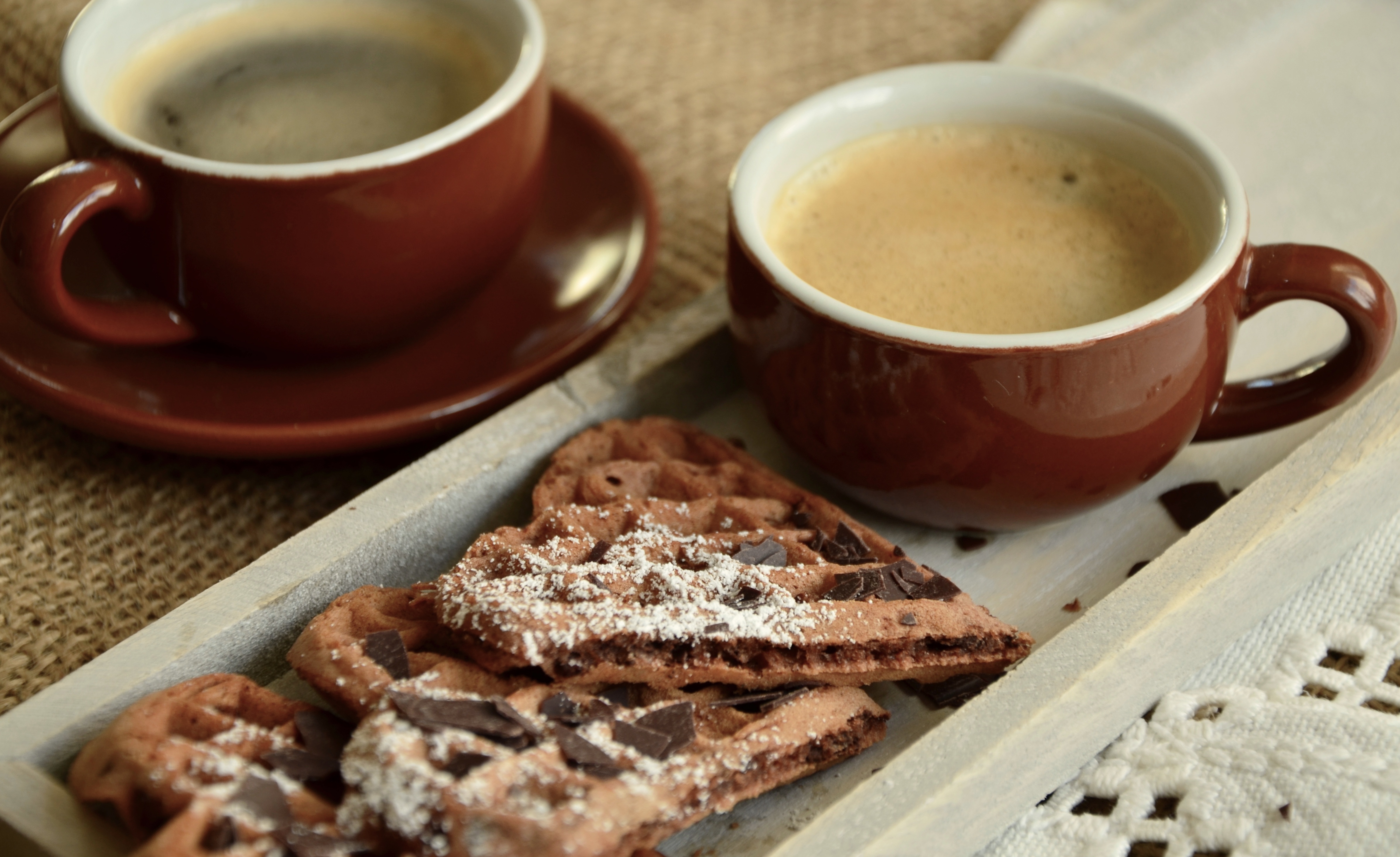 Auf einem Teller liegen mit Puderzucker bestäubte Waffelherzen. Zwei rote Kaffeetassen stehen drumherum.