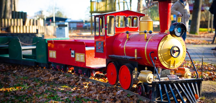 die rote Lokomotive auf dem Spielplatz für Kinder mit ihrem türkisenen und roten Zugteilen bei herbstlichem Wetter voller Blätter