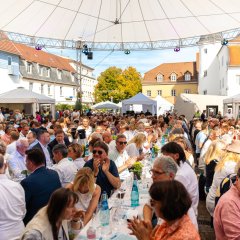 Auf dem Bild sind viele Menschen bei einer Weinprobe unter einem Festzelt zu sehen. Sie sitzen auf Bänken und trinken ihren Wein, während sie richtung Bühne schauen.