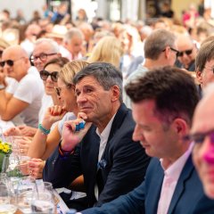 Auf dem Bild sind mehrere Männer zu erkennen, die nebeneinander auf der Bank sitzen und der Winzerfest Weinprobe zuhören.