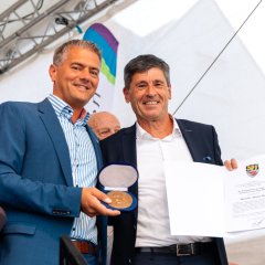 Auf dem Bild schauen Bürgermeister Steffen Jung und Dr. Markus Merk gemeinsam in die Kamera, während der Bürgermeister ihm eine goldene kleine Plakette in einem kleinen Kästchen überreicht - die Georg-Scheu-Plakette. In der anderen Hand hält Dr. Markus Merk eine Urkunde.