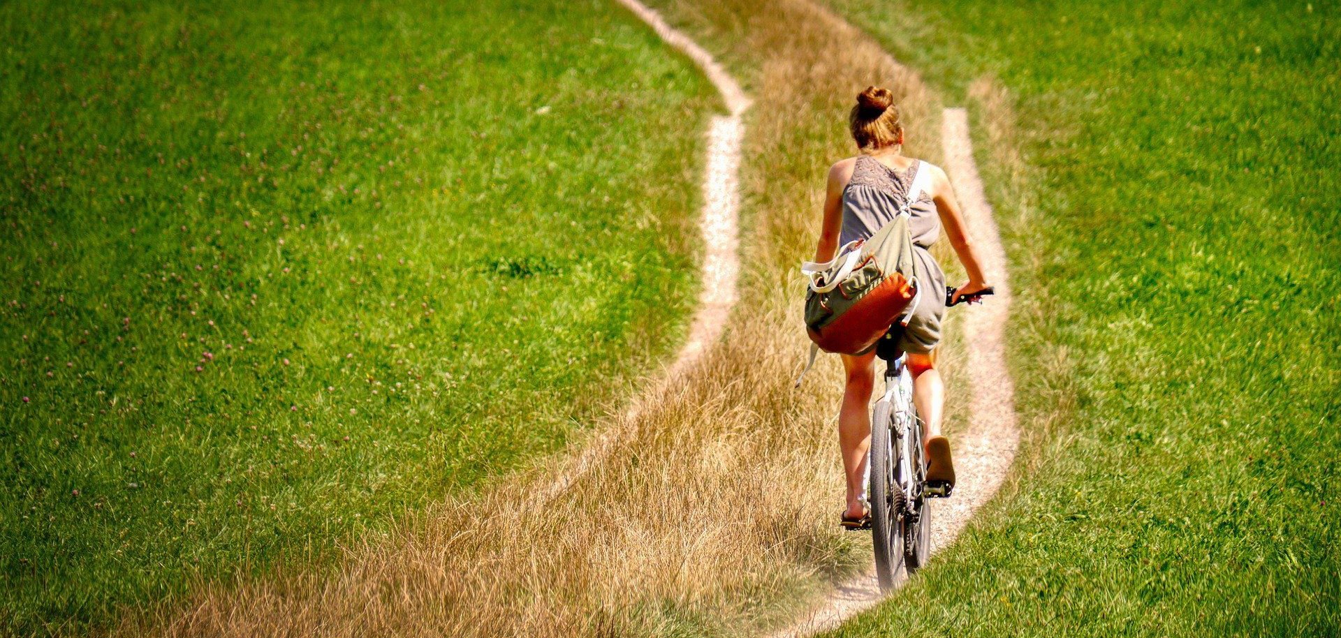 Auf dem Bild ist eine Fahrradfahrerin zu sehen, die auf einem Feldweg Fahrrad fährt. Rechts und links von ihr befinden sich grüne Felder.
