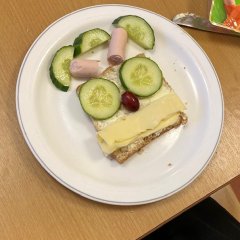 Auf dem Bild ist ein Brot zu sehen, was auf einem teller liegt. es ist belegt mit einer Scheibe Käse als Mund und Gurkenscheiben als Augen. Die Nase ist eine Traube. Oberhalb des Brotes stellt eine Wurst eine Art Krone dar.