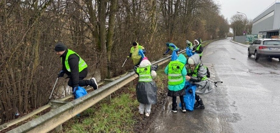 Man sieht Menschen und Kinder die entlang der Selz Müll bei Regenwetter sammeln. Die Personen tragen Warnwesten, Regenschutz und Müllbeutel während sie den Müll sammeln.