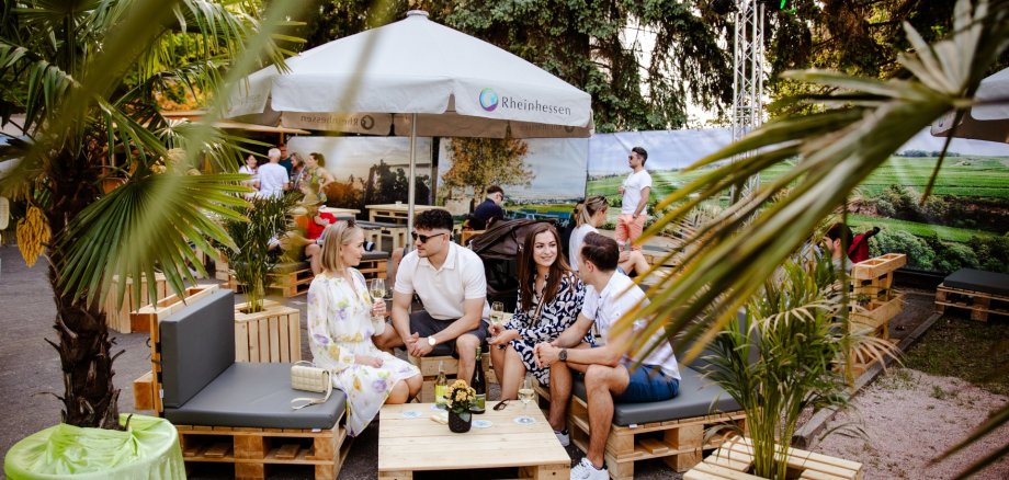 Auf dem Bild sieht man Menschen die sitzend das Weinfest "Scheutime" genießen und dabei Wein trinken und sich unterhalten, desweiteren sind ein Sonnenschirm, einige Palmen und Pflanzen zu sehen.