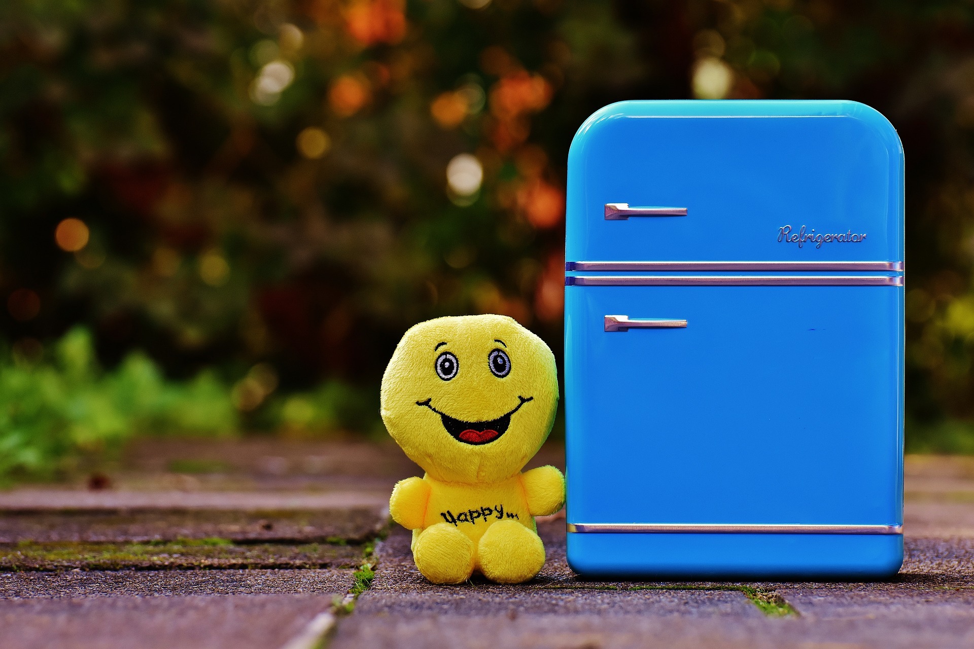 Man sieht eine kleine gelbe Stoffpuppe mit der Aufschrift "Happy" und einen kleinen blauen Kühlschrank.