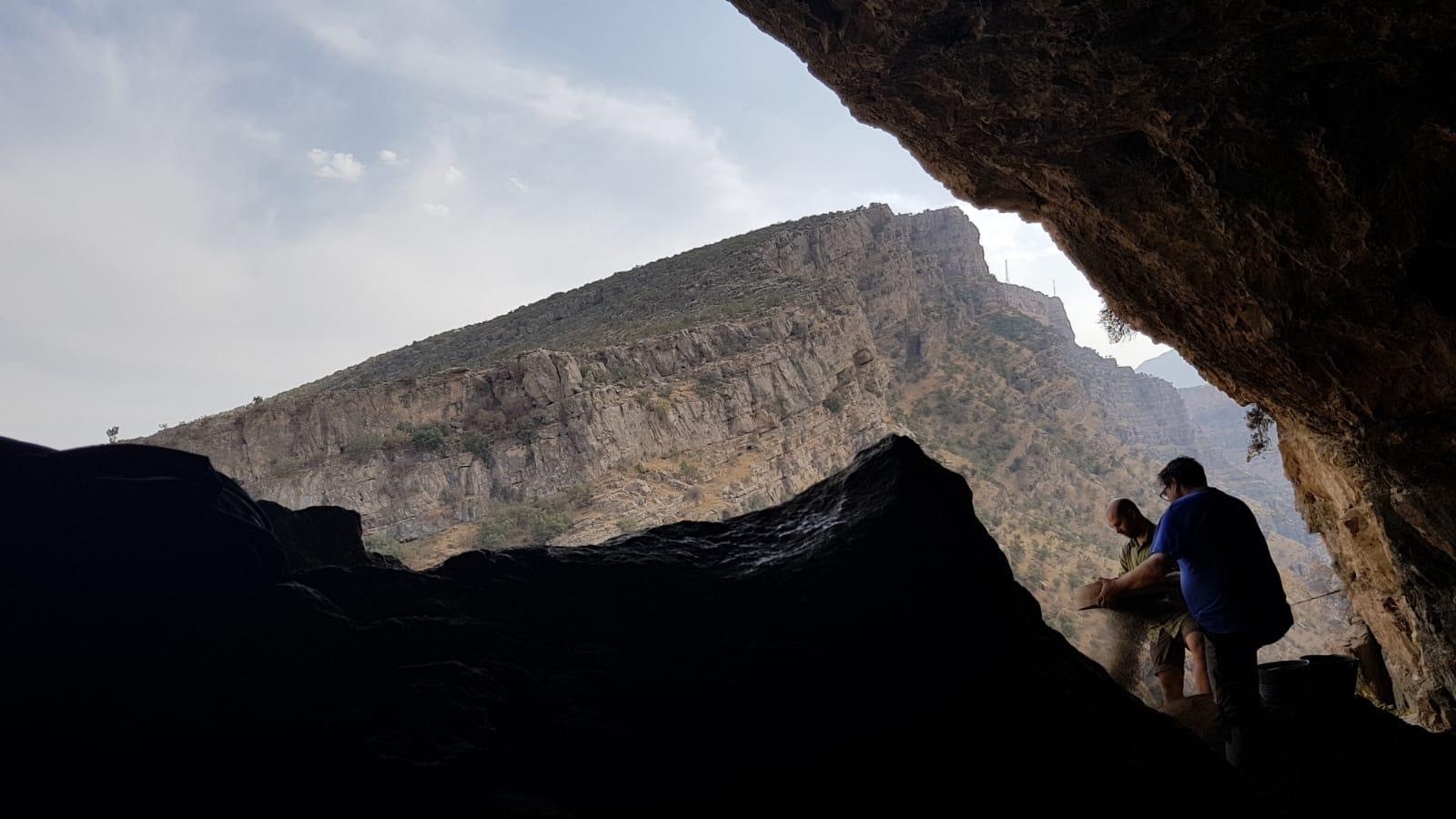 Das Bild zeigt eine Verbindung zwischen Mesopotamien, Anatolien und dem iranischen Hochland, man sieht eine kleine Ausgrabungsstätte und 2 Personen in einem Höhleneingang sowie einen Berg im Hintergrund.