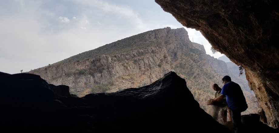 Das Bild zeigt eine Verbindung zwischen Mesopotamien, Anatolien und dem iranischen Hochland, man sieht eine kleine Ausgrabungsstätte und 2 Personen in einem Höhleneingang sowie einen Berg im Hintergrund.