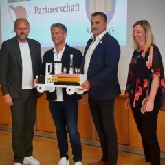 vier Personen stehen vor einer Kamera und halten zusammen eine Holzlokomotive in der Hand, in der Marillenschnaps steht