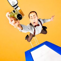 ein Mann in karriertem Hemd springt über einem Trampolin in der Luft und hält eine goldene Digitalkamera in die Kamera, dabei schaut er sehr erstaunt mit aufgerissenen Augen