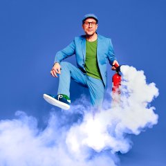 ein Mann in blauem Anzug und grünem Shirt darunter hält in seiner linken Hand einen Feuerlöscher, der grade auslöst und eine Schaumwolke verursacht