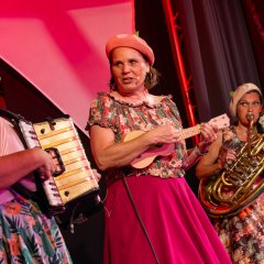 drei Frauen stehen auf einer Bühne und tragen bunte Kostüme, während jede von ihnen ein Musikinstrument in der Hand hält
