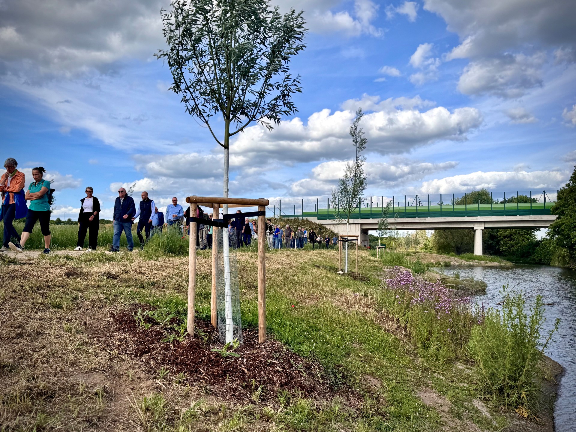 rechts im Bild ist ein Fluss abgebildet, links daneben läuft eine Gruppe von Menschen auf einem Feldweg