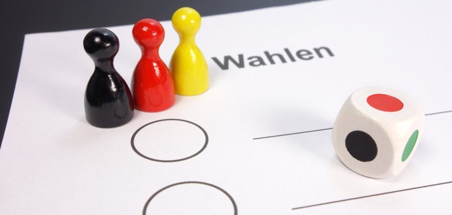 Man sieht einen Wahlen Zettel mit 3 Spielfiguren in den Farben Schwarz-Rot-Gold/Gelb und einen Würfel.