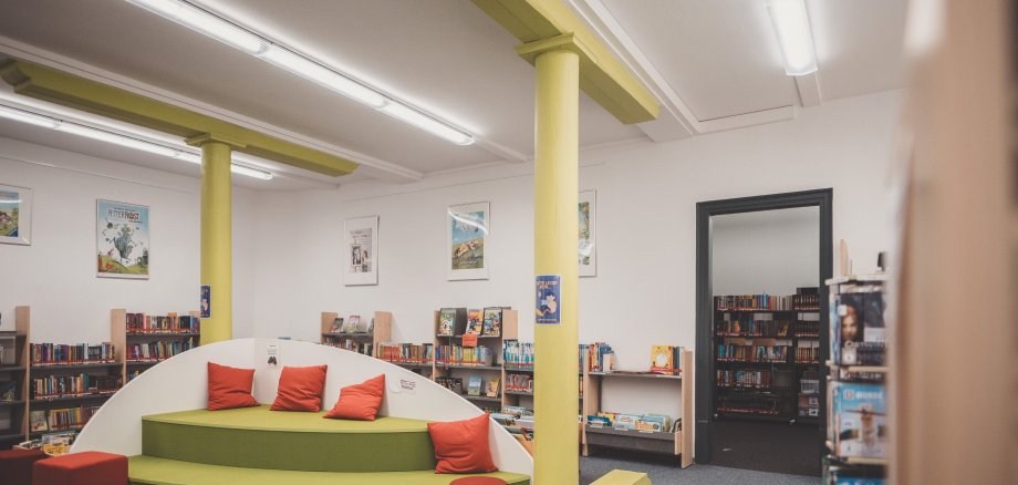eine Bücherei von innen mit einem Sitzbereich in den Farben orange und grün, im Hintergrund sind Bücherregale, gefüllt mit vielen Büchern zu sehen