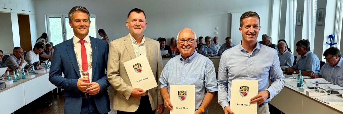 Vier Personen stehen mit Urkunden in der Hand in einem Sitzungssaal voller Menschen.
