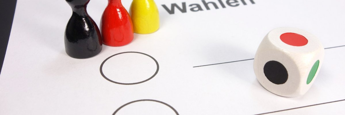 Drei Mensch ärgere Dich nicht Figuren in Schwarz Rot Gelb stehen mit einem Würfel auf einem Wahlzettel