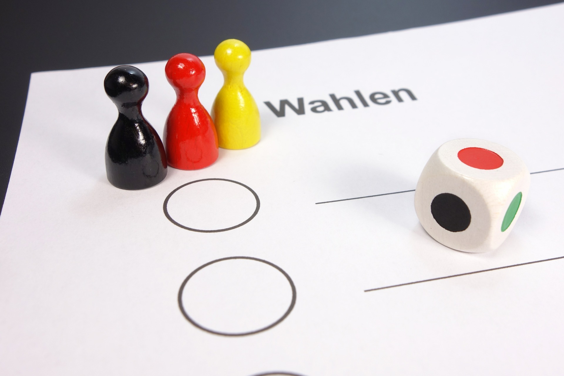 Drei Mensch ärgere Dich nicht Figuren in Schwarz Rot Gelb stehen mit einem Würfel auf einem Wahlzettel