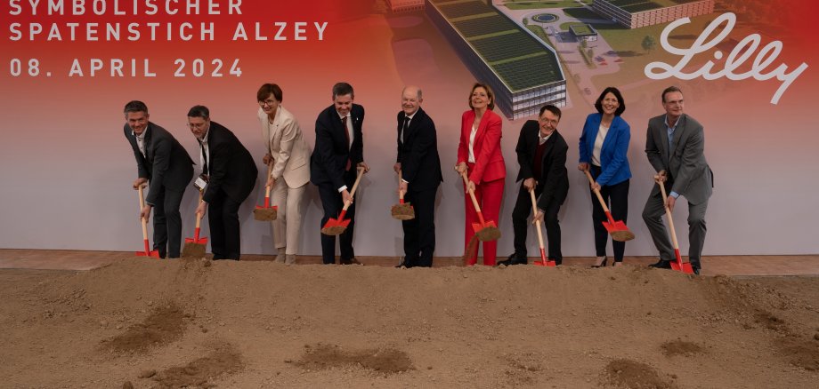 Neun Personen stehen mit Spaten in den Händen vor einer Stellwand, die das neue Firmengebäude von Eli Lilly in Alzey zeigt.