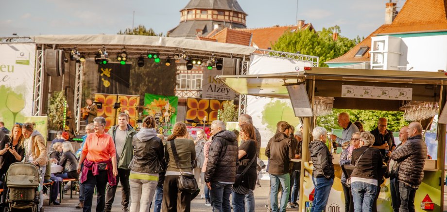 Viele Menschen stehen vor einer Bühne in kleinen Gruppen zusammen. Es wird gelacht, mit Weingläsern angestoßen und sich unterhalten.