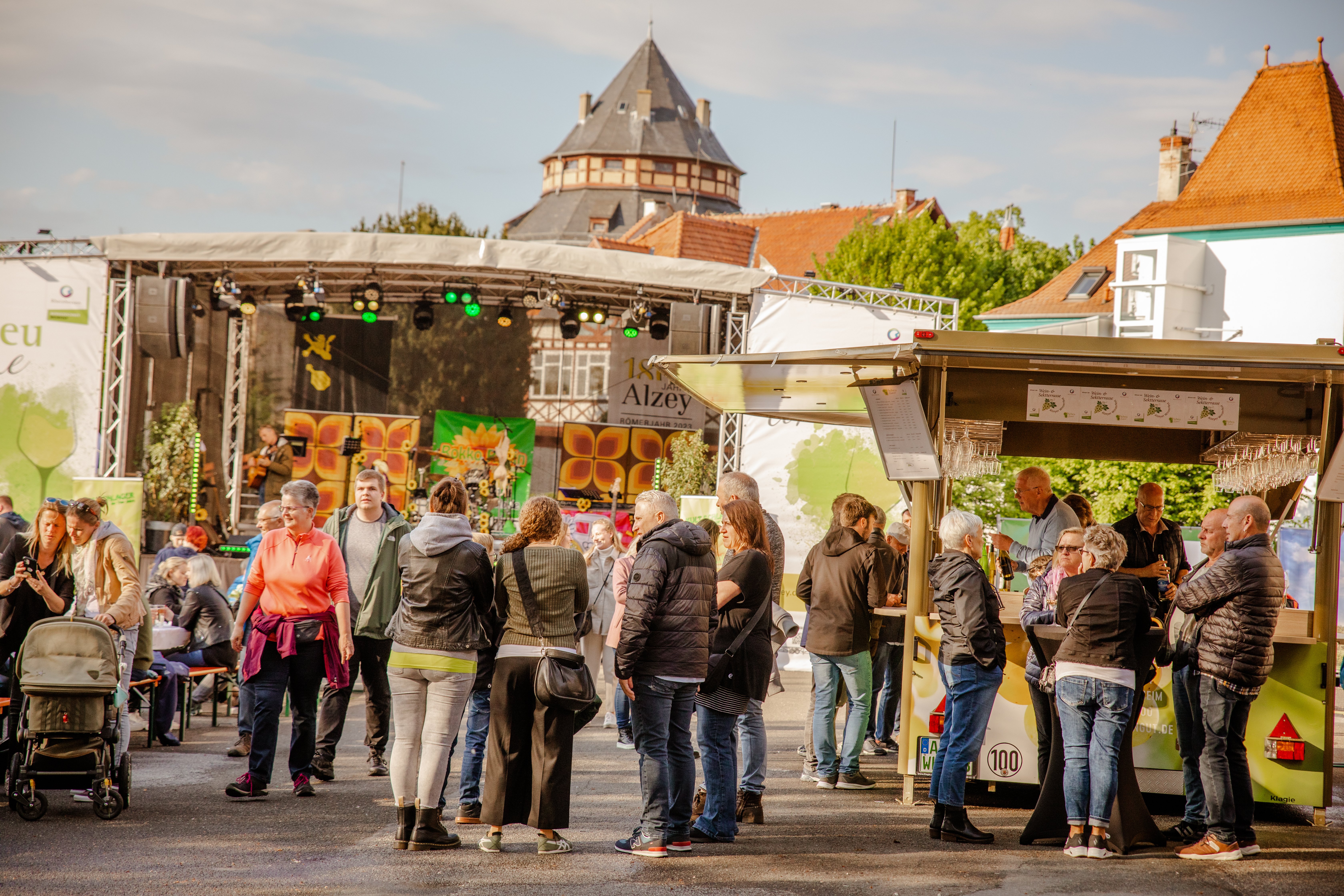 Viele Menschen stehen vor einer Bühne in kleinen Gruppen zusammen. Es wird gelacht, mit Weingläsern angestoßen und sich unterhalten.