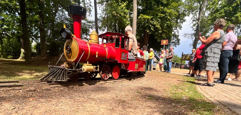 Eine kleine rote Lokomotive fährt die Kinder. Am Bahnsteig stehen viele Erwachsene und freuen sich mit den Fahrgästen.