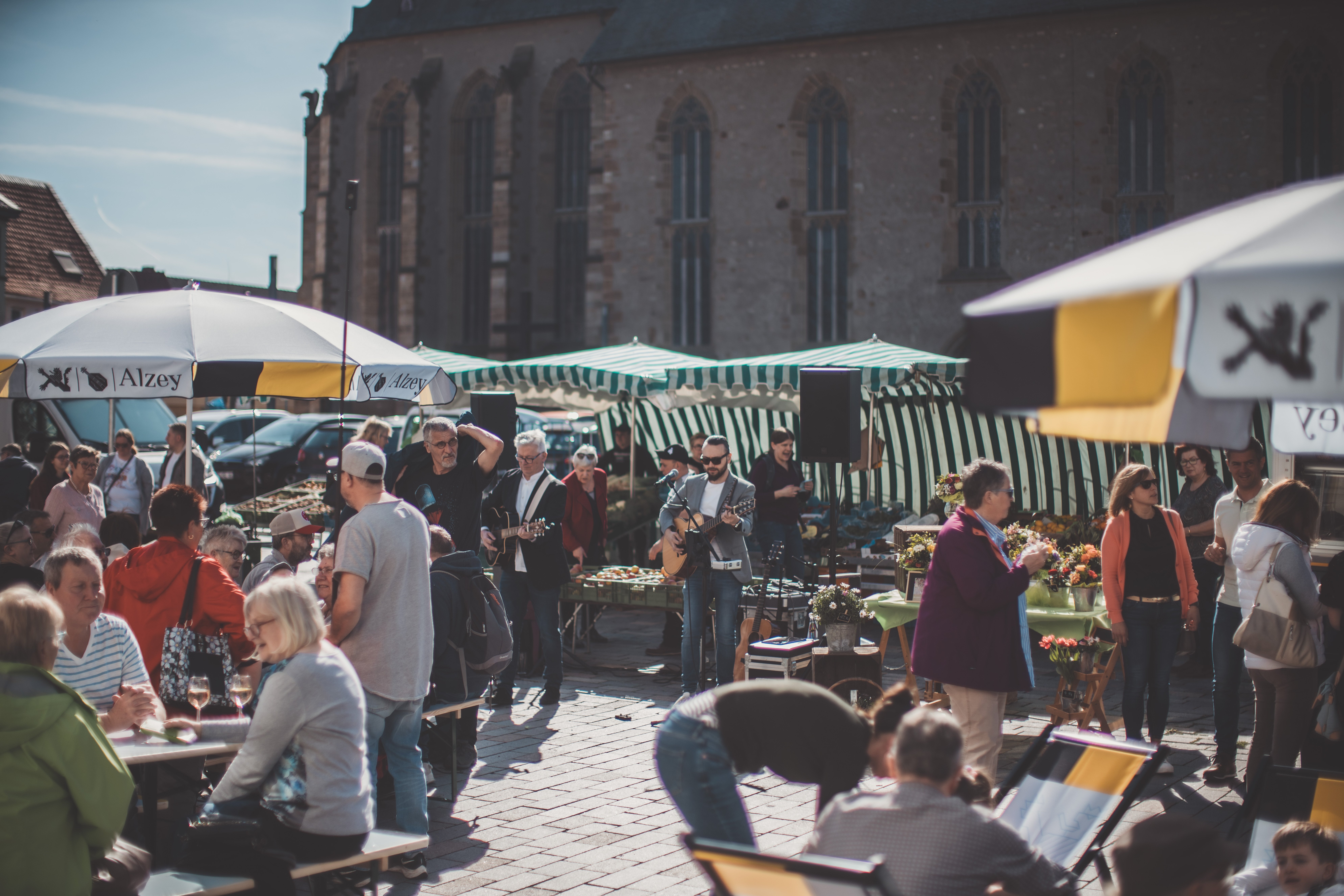 Zwischen den Marktständen sitzen die Leute unter Schirmen auf Bierzeltgarnituren. Zwei Musiker mit Gitarren und Sonnenbrillen stehen in der Mitte.
