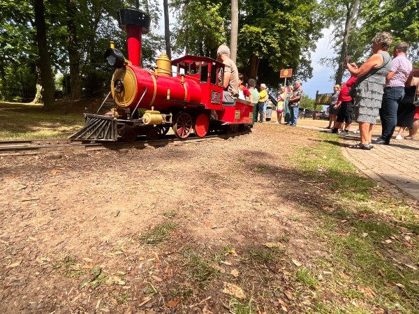 Eine kleine rote Lokomotive fährt die Kinder. Am Bahnsteig stehen viele Erwachsene und freuen sich mit den Fahrgästen.