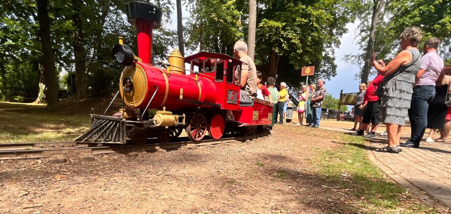 Eine kleine rote Lokomotive fährt die Kinder. Am Bahnsteig stehen viele Erwachsene und freuen sich mit den Fahrgästen.
