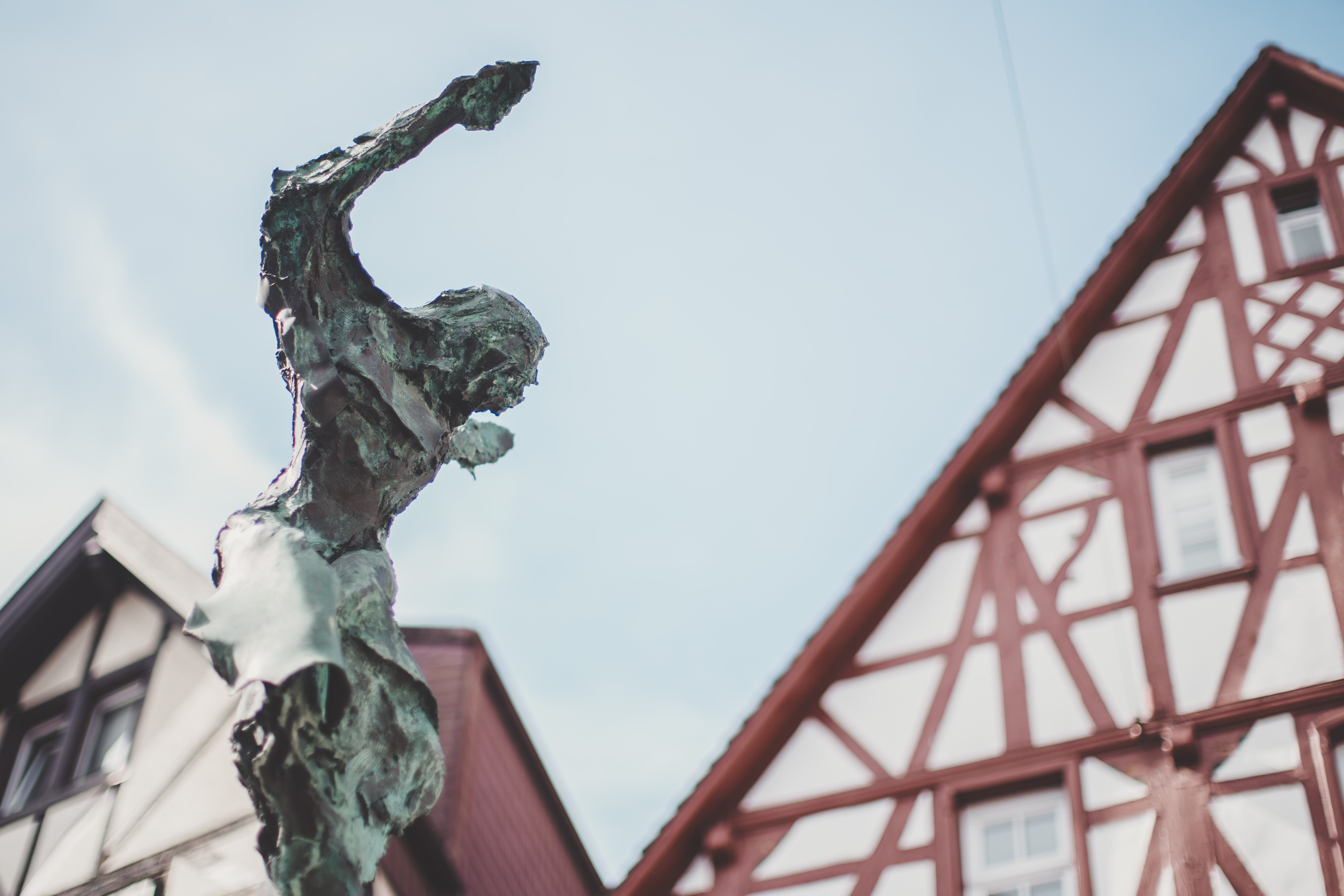 Eine Bronzeskulptur. Im Hintergrund ist ein weiß-rotes Fachwerkgebäude und blauer Himmel zu sehen.