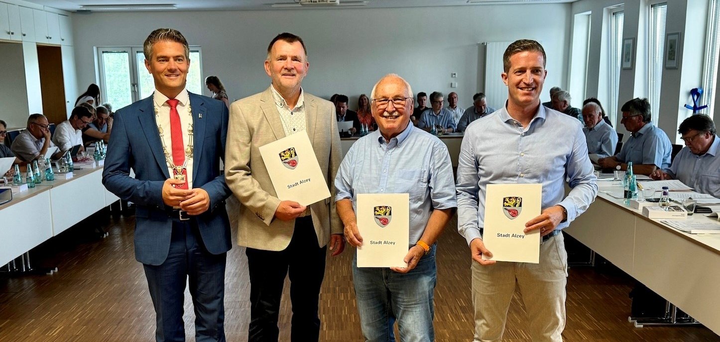 Vier Personen stehen mit Urkunden in der Hand in einem Sitzungssaal voller Menschen.