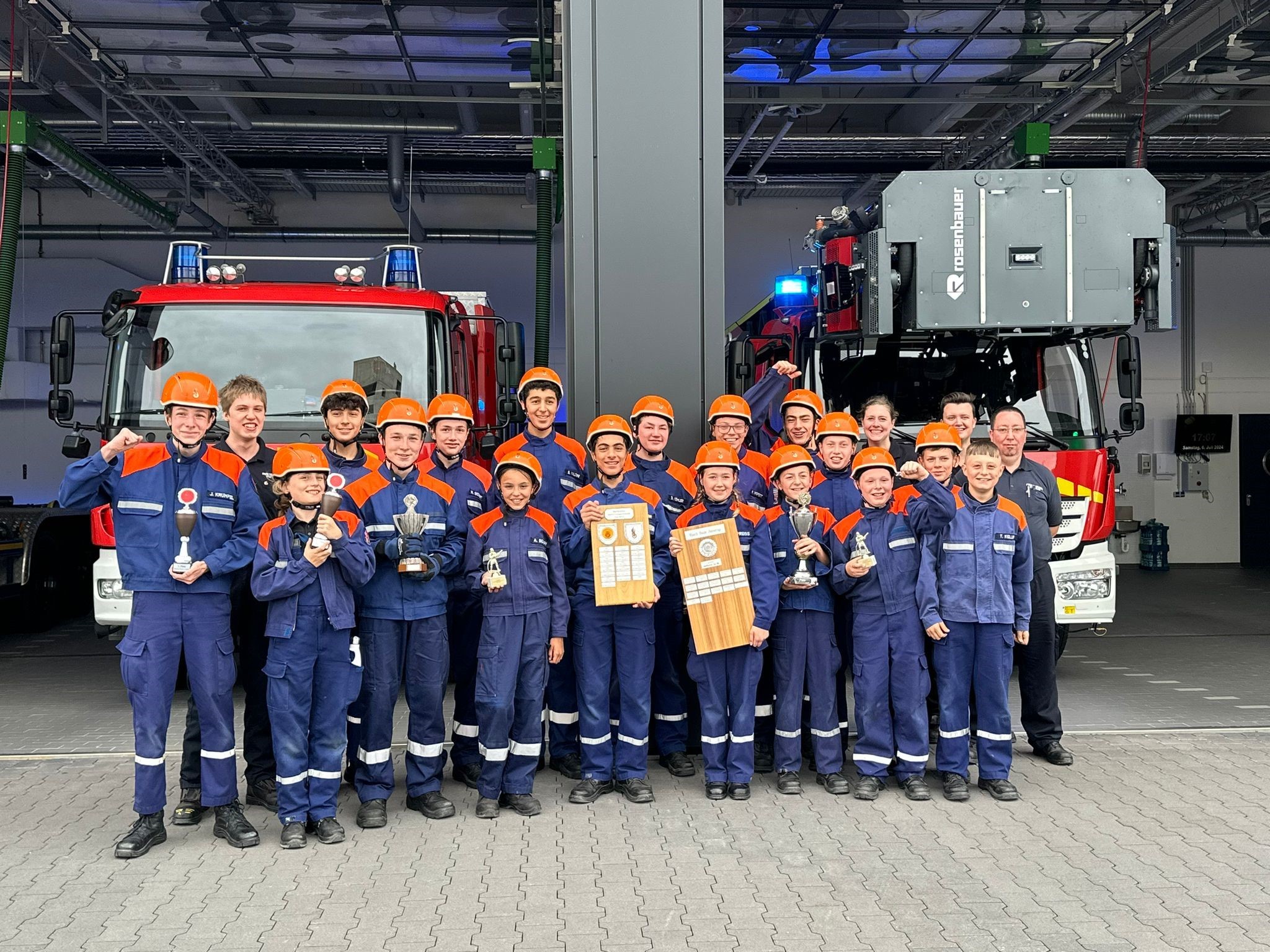 Eine große Gruppe von Kindern und Jugendlichen stehen in ihren blau-orangenen Uniformen vor zwei Einsatzfahrzeugen und zeigen stolz ihre gewonnen Pokale.