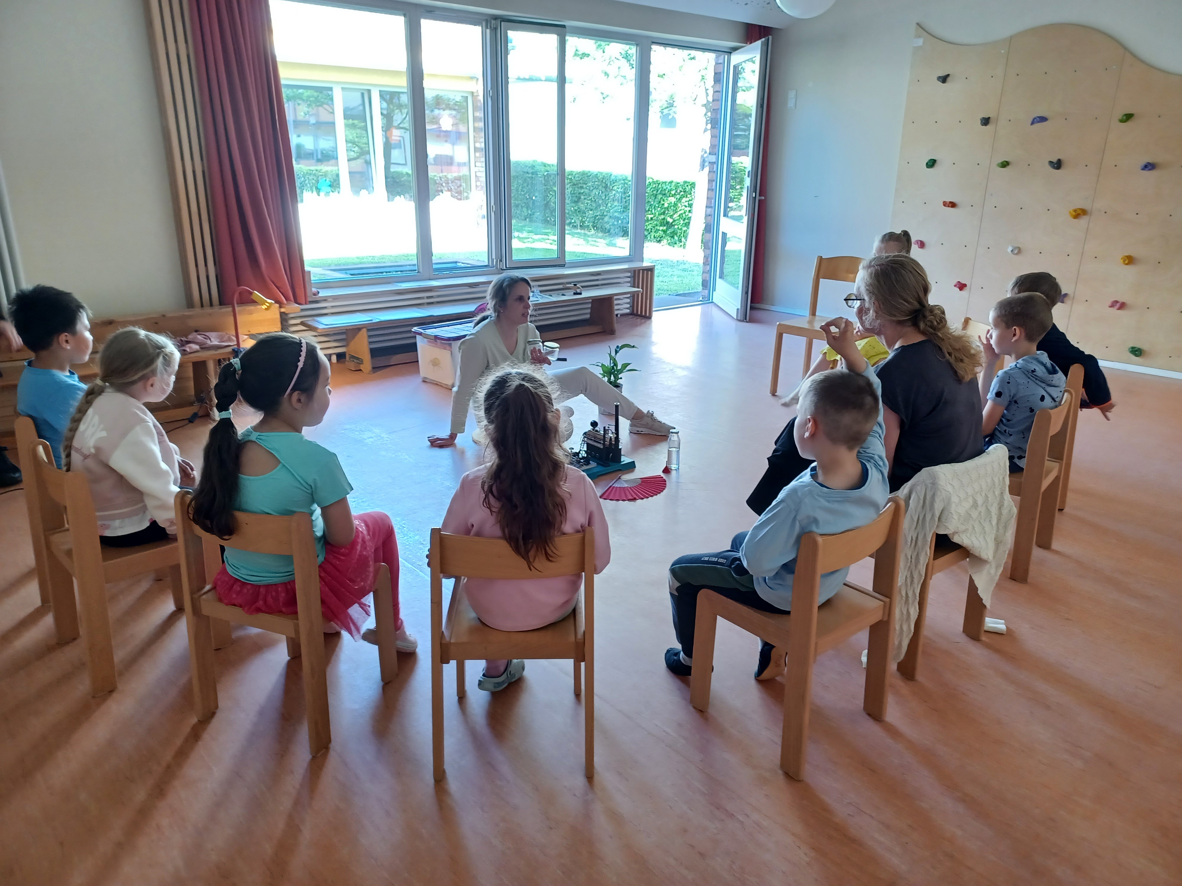 In einem Stuhlkreis sitzen die Kinder und beobachten die Frau in der Mitte dabei, wie sie eine kleine Dampfmaschine anwirft.